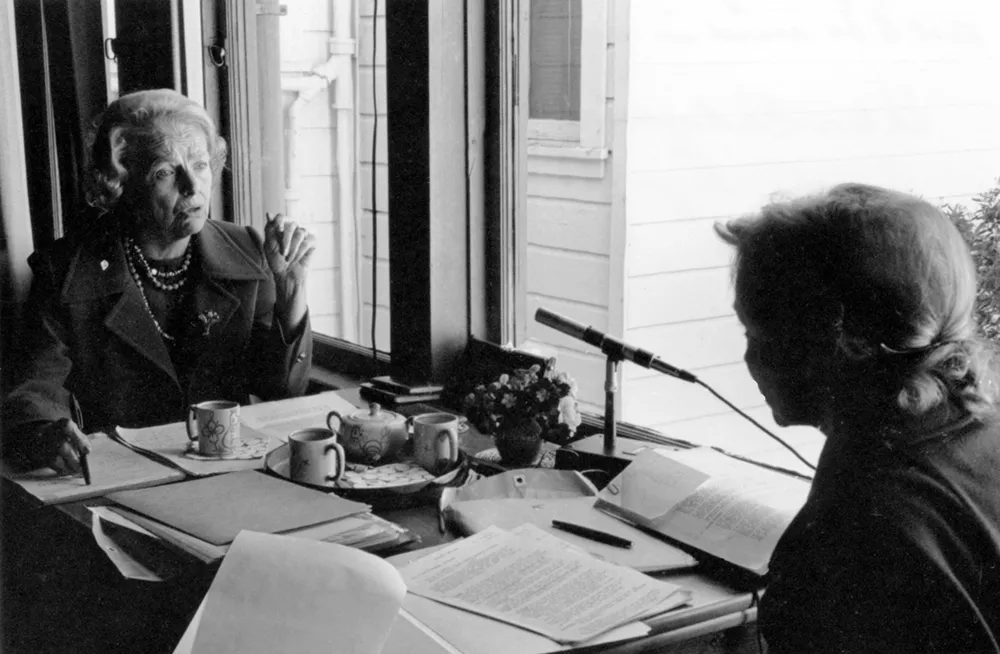 Amelia Fry interviews Helen Gahagan; both are seated at a table piled with papers, teacups, and a microphone.